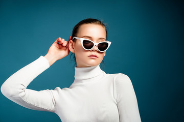 Portrait of a beautiful young girl in white knitted golf and sunglasses on a blue background