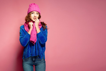 Modern young woman wearing blue sweater and pink hat, and scarf posing, making funny facial expression. Retouched, studio portrait.