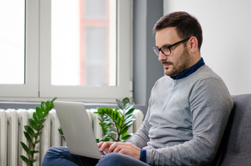 Man sitting on sofa at home and working on laptop, side view