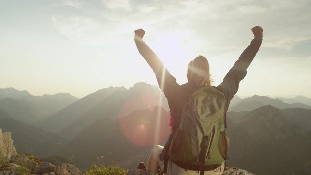SLOW MOTION, LENS FLARE, CLOSE UP: Unrecognizable female hiker sits on a rock and outstretches her arms as she observes the breathtaking Julian Alps. Cinematic shot of trekker girl reaching the summit