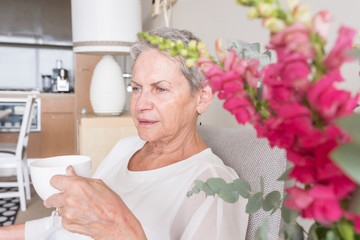 Beautiful senior woman with grey hair drinking tea in armchair with view of flowers and kitchen (selective focus)