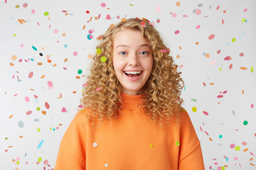 Obraz na płótnie Canvas Celebrating happiness, young woman with big smile stands under the falling confetti.Curly blonde in orange sweater enjoying life moment, came to the holiday.