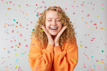 Portrait of a cheerful beautiful girl wearing orange sweater keeps palms near face standing...