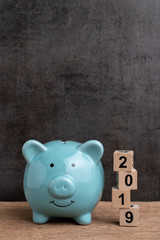 Year 2019 happy saving target, budget, investment or finance goals concept, blue piggy bank and stack of cube wooden block building unstable risk year number 2019 on wood table with dark background