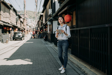 Obraz premium full length of young girl backpacker visiting Hanamikoji street standing on road holding paper map. japanese wooden building house hanging bamboo curtain on roof. woman finding direction destination.