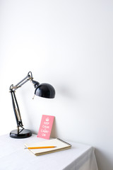 Retro black desk lamp white blank notepad and a yellow pencil and a motivational quote poster on white table top on white wall background with copy space product mock up placement