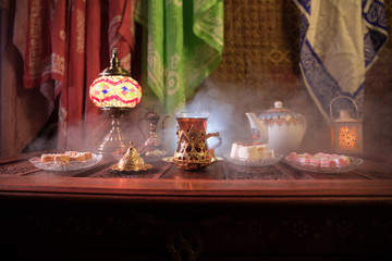 Arabian tea in glass with eastern snacks on vintage wooden surface. Eastern tea concept. Low light lounge interior with carpet. Empty space.