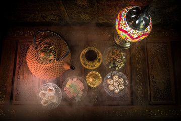 Arabian tea in glass with eastern snacks on vintage wooden surface. Eastern tea concept. Low light lounge interior with carpet. Empty space.