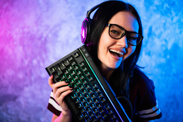 Nerd geek young adult women holding gaming keyboard over colorful pink and blue neon lit wall....