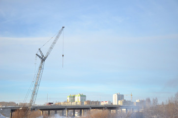 Gray tower crane towering over the bridge on the background of high-rise buildings and blue sky