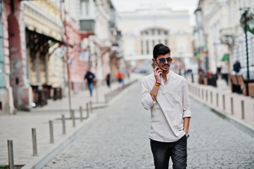 Stylish indian model man in casual clothes and sunglasses posed outdoor at street of India and speaking on mobile phone.