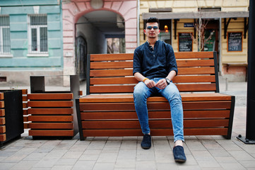 Stylish indian model man in casual clothes, black shirt and sunglasses posed outdoor at street of India and sitting on bench.