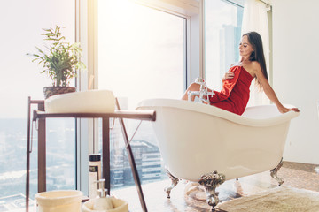 Slim young woman in towel sitting on bathtub in luxurious bathroom
