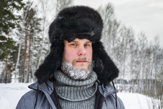 Siberian Russian man with a beard in hoarfrost in freezing cold in the winter freezes in a village in a snowdrift and wears a hat with a earflap.