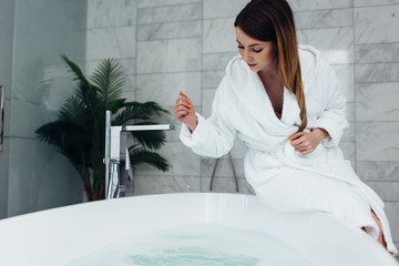 Pretty slim woman wearing bathrobe sitting on edge of bathtub filling up with water