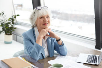Stylish mature middle aged woman writer with gray hair and wrinkles looking and smiling happily at camera, clasping hands, being in good mood, feeling inspired while working on her new book