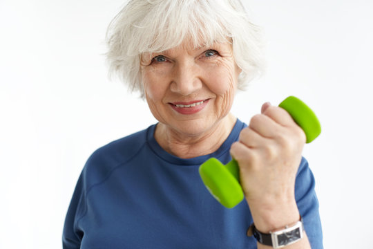 Close up image of energetic sporty mature woman with gray hair and wrinkles exercising indoors, doing bicep curls, holding green dumbbell and smiling happily at camera. Sports, age and fitness