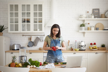 Beautiful young brunette female in denim jumpsuit cooking dinner in modern light kitchen interior, peeling red bell pepper, making healthy salad, watching video recipe online on portable computer