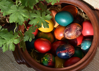 Pot full of multicolored Easter eggs. Green leaves of geranium. Rustic earthenware. Handmade rug at background.