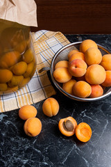 Fresh organic harvested apricots in steel colander and big glass jar with canned apricot compote on dark marble..