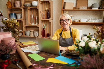 Portrait of smiling female small business owner using laptop and looking at camera in flower shop,...