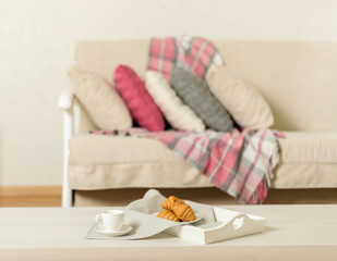 Fototapeta na wymiar Beige sofa with plaid, colorful pillows (pink, grey, white) and coffee with croissantsin the living room.