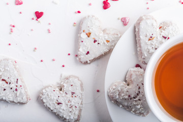 Fototapeta na wymiar White tea cup with tea and Valentine's day white coconut heart shaped cookies with red and pink heart sprinkles. Copy space