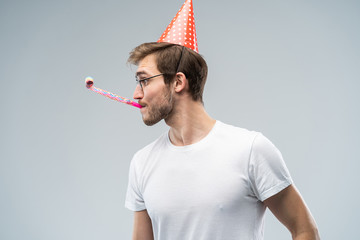 Studio shot of unshaven young Caucasian male blowing whistle while celebrating birthday, having...