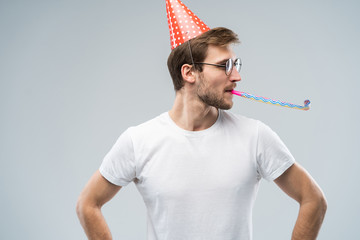 Studio shot of unshaven young Caucasian male blowing whistle while celebrating birthday, having...