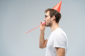 Studio shot of unshaven young Caucasian male blowing whistle while celebrating birthday, having...