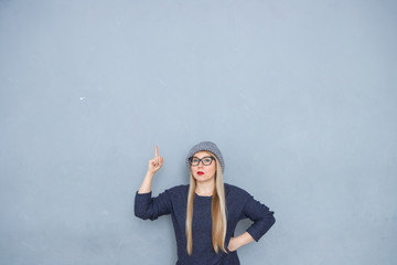 A blonde girl wearing glasses and red lips, dressed in casual style and wearing a hat, stands on a blue background and points her hands to the top.