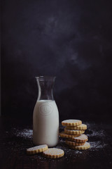 A bottle of milk and a stack of cookies sprinkled with icing sugar, against a dark background