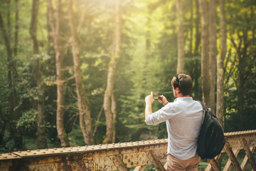 a dark-haired man on a bridge over a river and photographing the lush and vibrant nature in Georgia with a new phone. an idea for advertising a gadget and things, a tourist with a black backpack