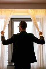Businessman in dark hotel room opens curtains on window to the morning light