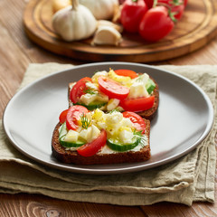 Smorrebrod - traditional Danish sandwiches. Black rye bread with boiled egg, cream cheese, cucumber, tomatoes on dark brown wooden background, side view