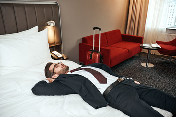 Businessman in business suit sitting with smartphone on bed at hotel room