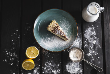 Photo of chocolate cheescake with lemon, milk, caster sugar and sieve on wooden background. Dark food Photography of sweet and illuminated dessert from above.