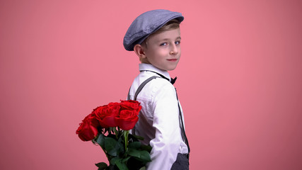 Little gentleman kid hiding roses behind back and turning to camera, smiling