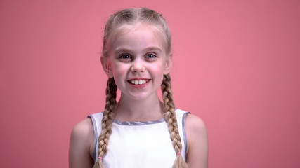 Pretty female kid with braids smiling on camera against pink background, joy