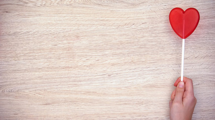 Hand holding heart-shaped lollipop against wooden background, love, top view