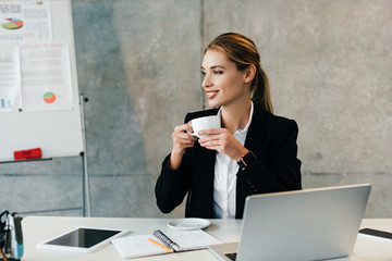 pretty smiling businesswoman sitting at workplace and drinking coffee