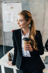selective focus of smiling businesswoman staying by work desk with disposal cup of coffee