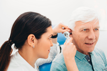 doctor examining elderly patient ear , using otoscope, in doctors office. Senior man getting medical ear exam at clinic.