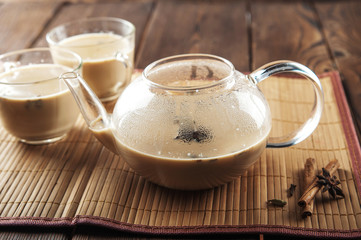 Masala tea on a textural wooden background. A teapot is a transparent tea pot with mugs and Indian national tea masala. Milk, ginger, cardamom, cloves, nutmeg, star anise, cinnamon