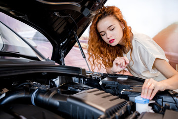 Young beautiful red-haired girl standing in front of an emergency car with an open car hood trying to fix the fault