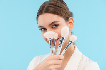 Beautiful young woman posing isolated over blue background holding makeup brushes.