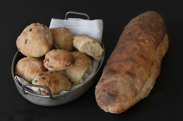 Just baked sourdough homemade bread and buns with seeds - rustic quality. Buns lying in old antique metal basket with napkin. Beautiful picturesque image on black background.