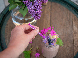Woman’s hand holding straw in jar with smoothie made from berries and banana. Lilac on background.