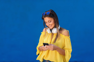 Smiling cute teenage girl in yellow blouse with sunglasses and earpieces around neck using smart phone while standing in front of blue background.