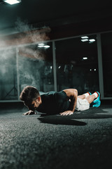 Muscular Caucasian bodybuilder doing jumping push ups in dark. Dust in the air, gym interior,...